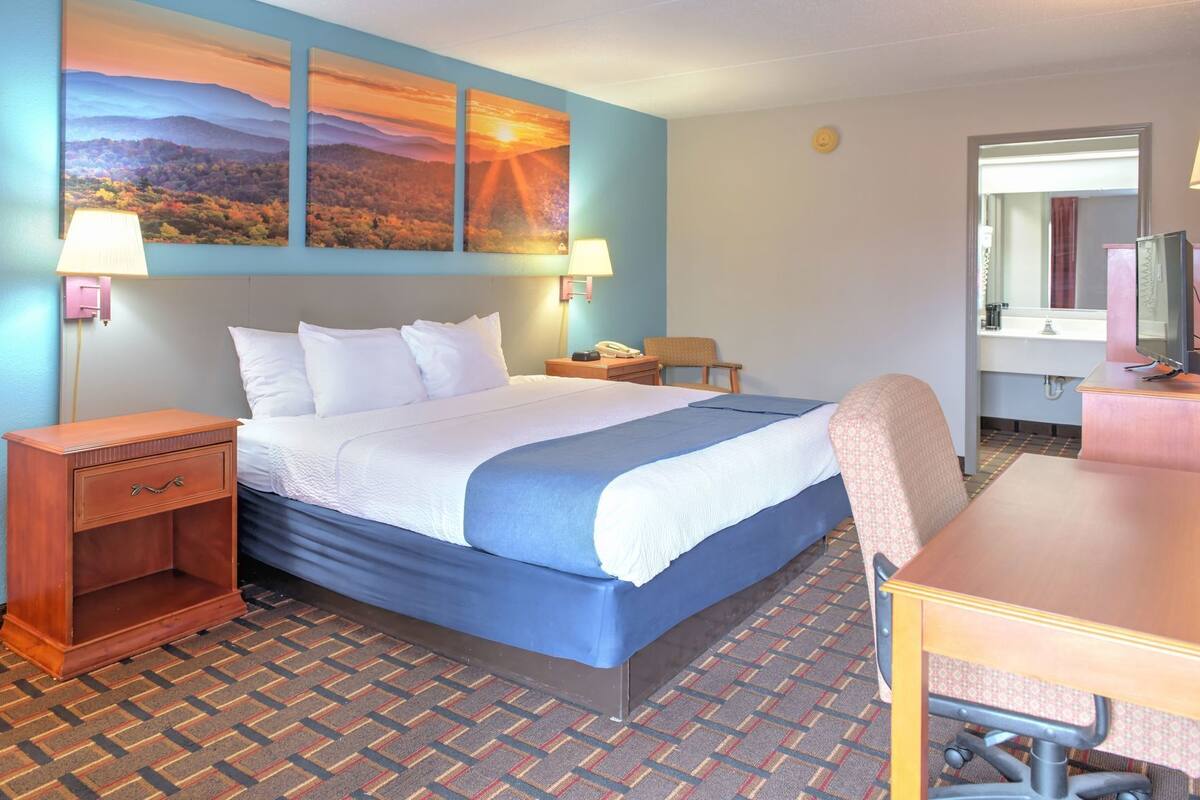 A hotel room with a bed, bedside tables, a desk, a chair, a television, and a painting on the wall.