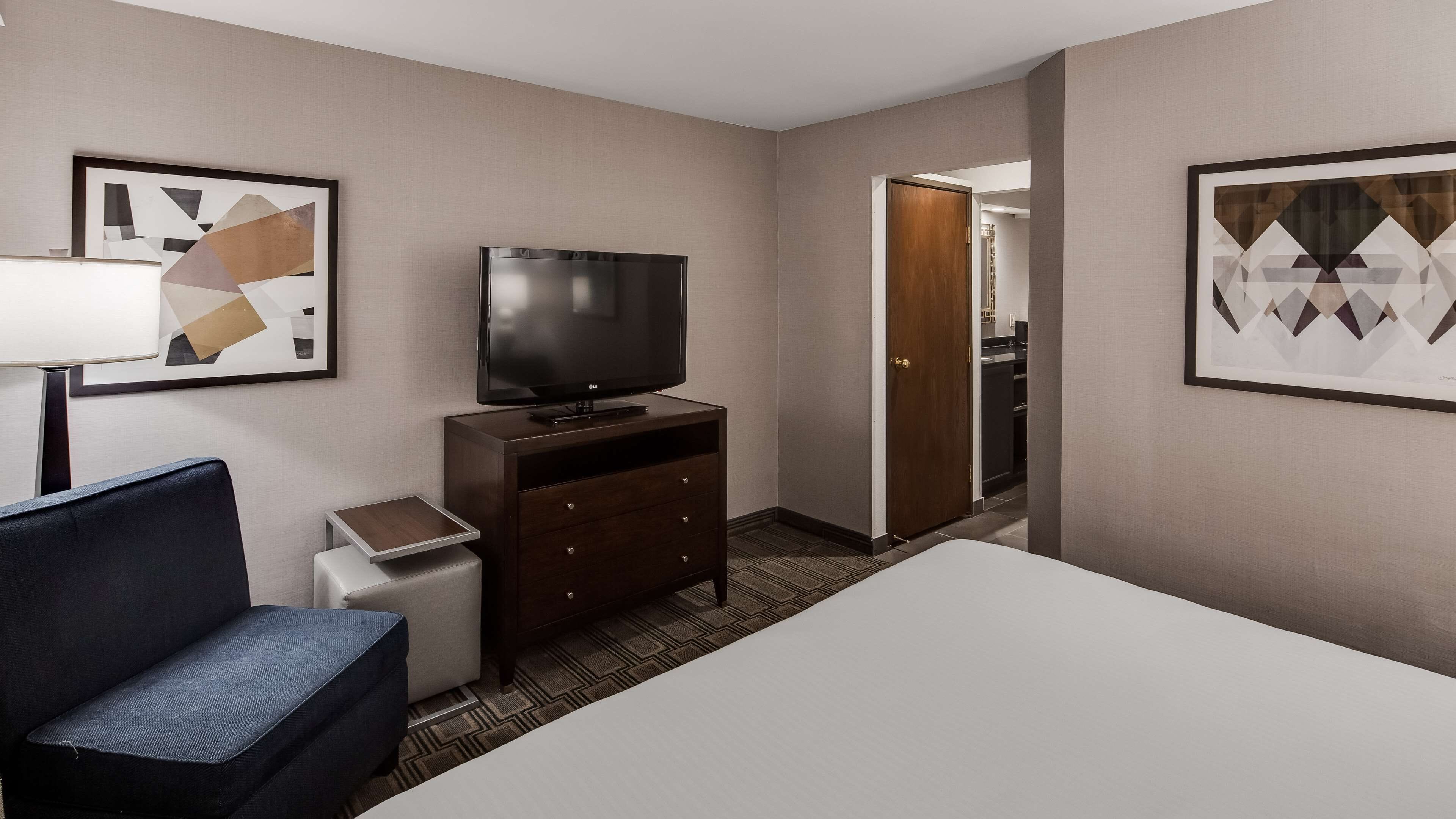 Una habitación de hotel con cama, televisor sobre una cómoda de madera, una silla y obras de arte en las paredes.