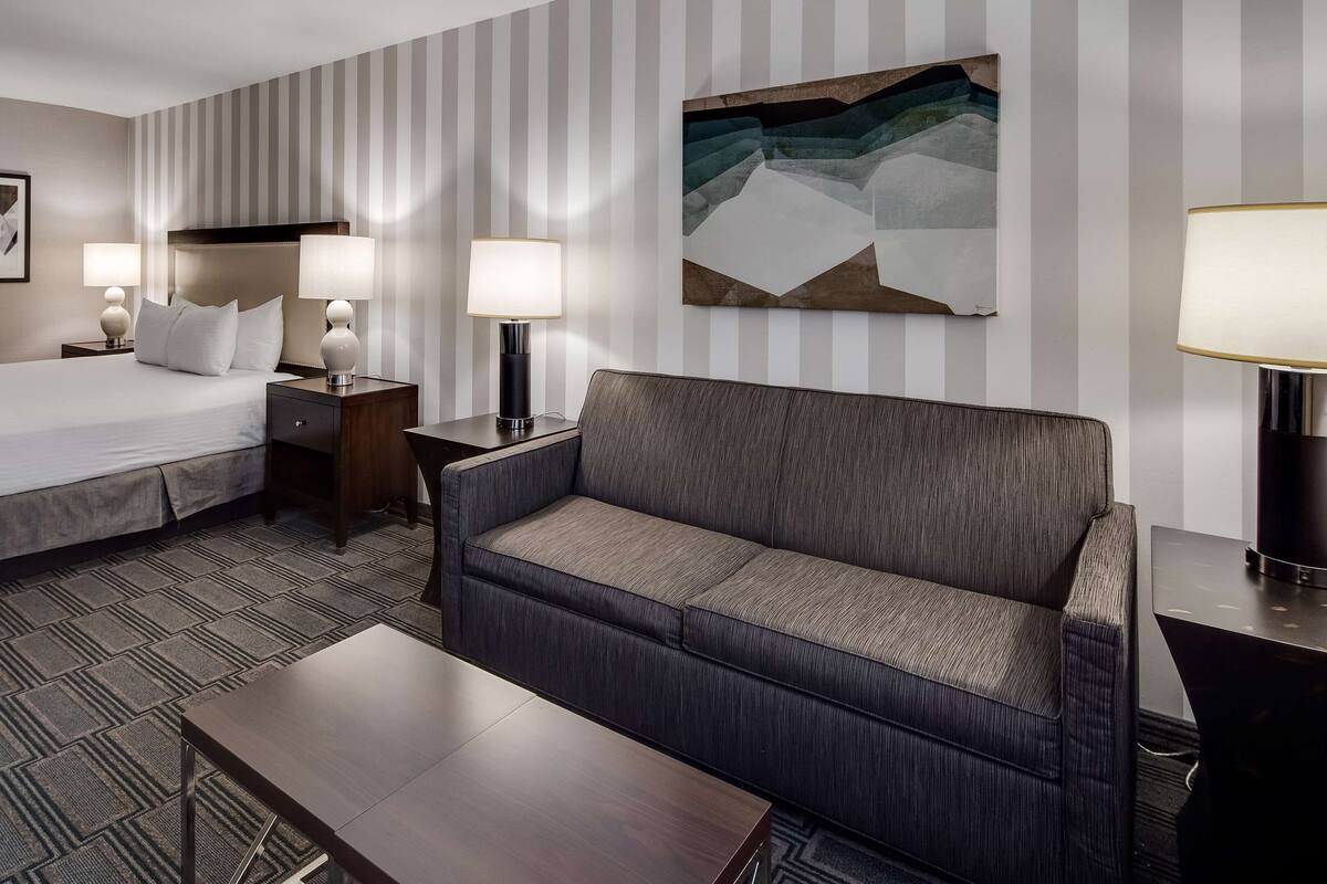 A hotel room with a bed, a sofa, a nightstand, a lamp, and a painting on the wall.