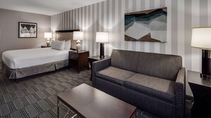 A hotel room with a bed, a sofa, a nightstand, a lamp, and a painting on the wall.