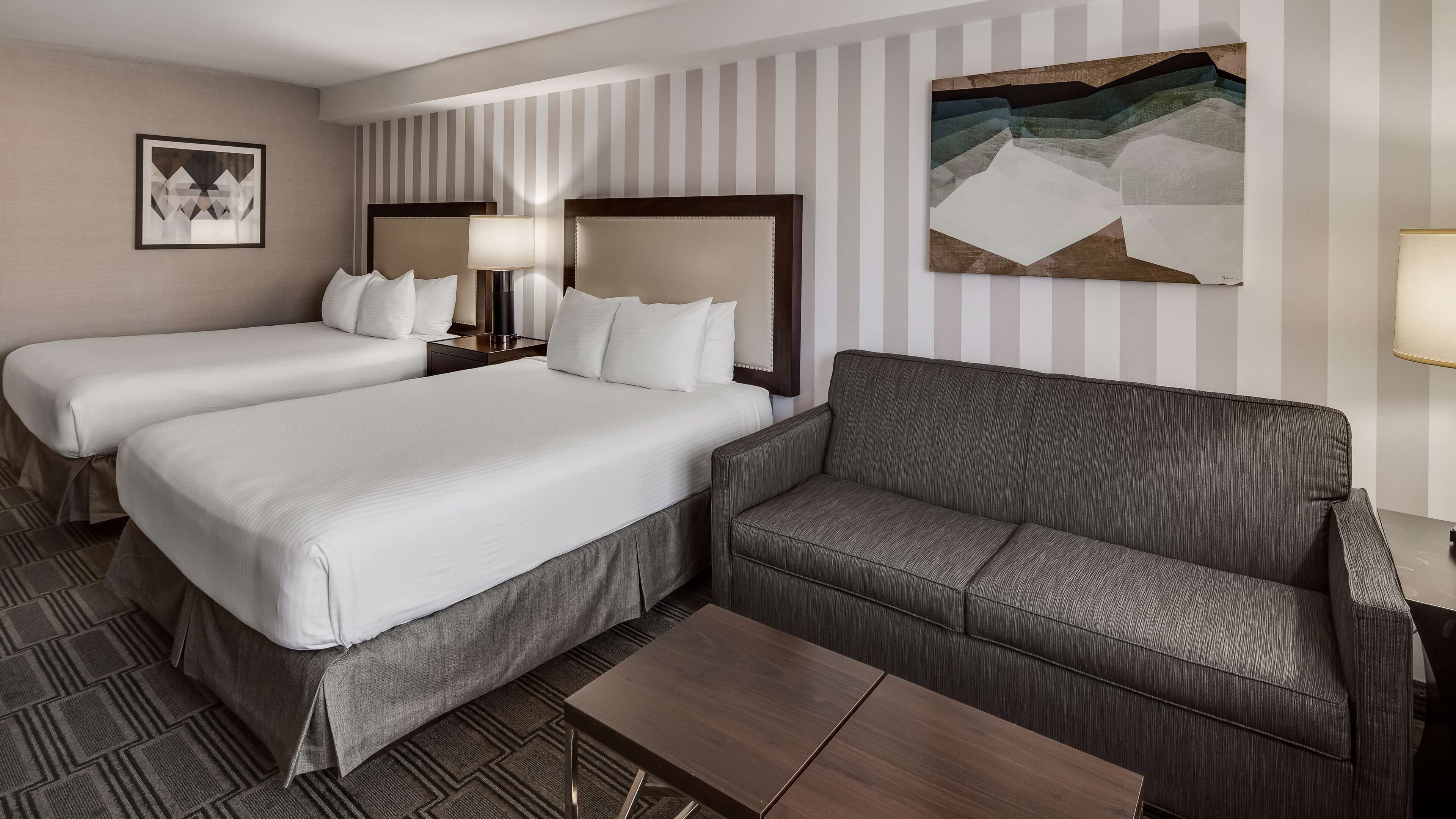 Habitació d'hotel amb dos llits, un sofà gris, una taula de fusta petita i paper pintat a ratlles.