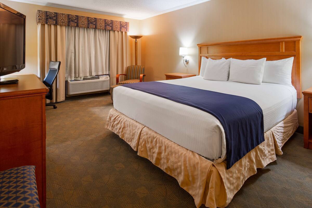 A hotel room with a large bed, a desk with a flat-screen TV, an air conditioning unit, and a bedside lamp.