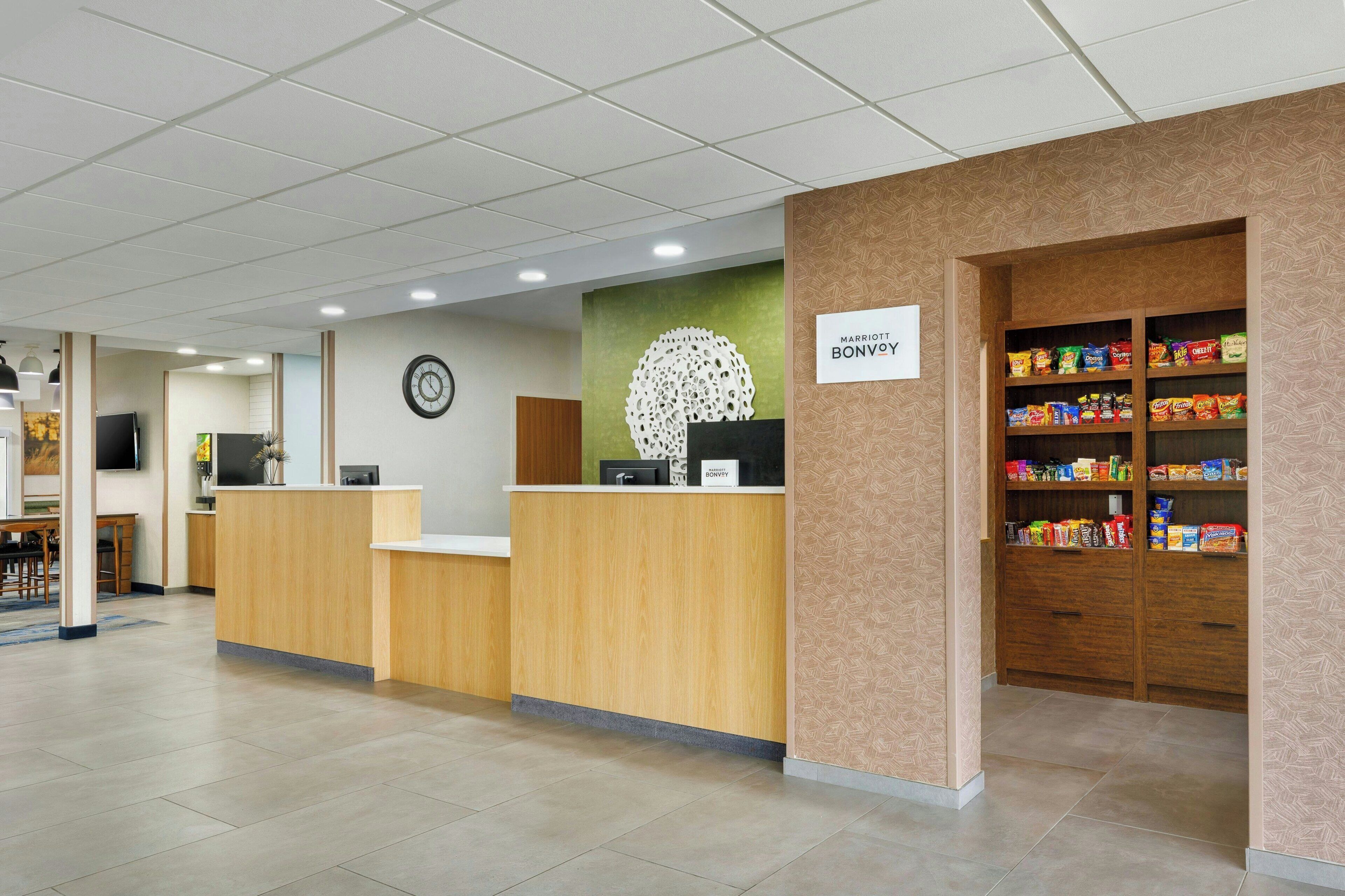 A hotel reception area with a wooden reception desk, a clock on the wall, a sign for Marriott Bonvoy, and a snack shelf.