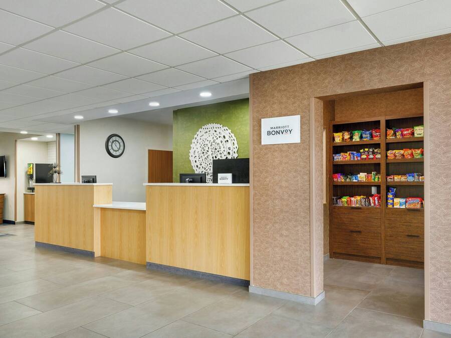 A hotel reception area with a wooden reception desk, a clock on the wall, a sign for Marriott Bonvoy, and a snack shelf.