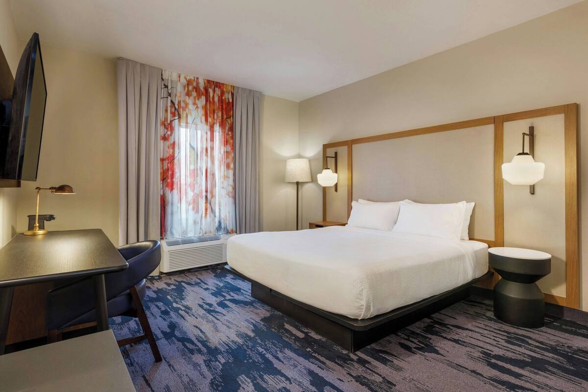 A hotel room with a bed, desk, chair, and a large window with curtains.