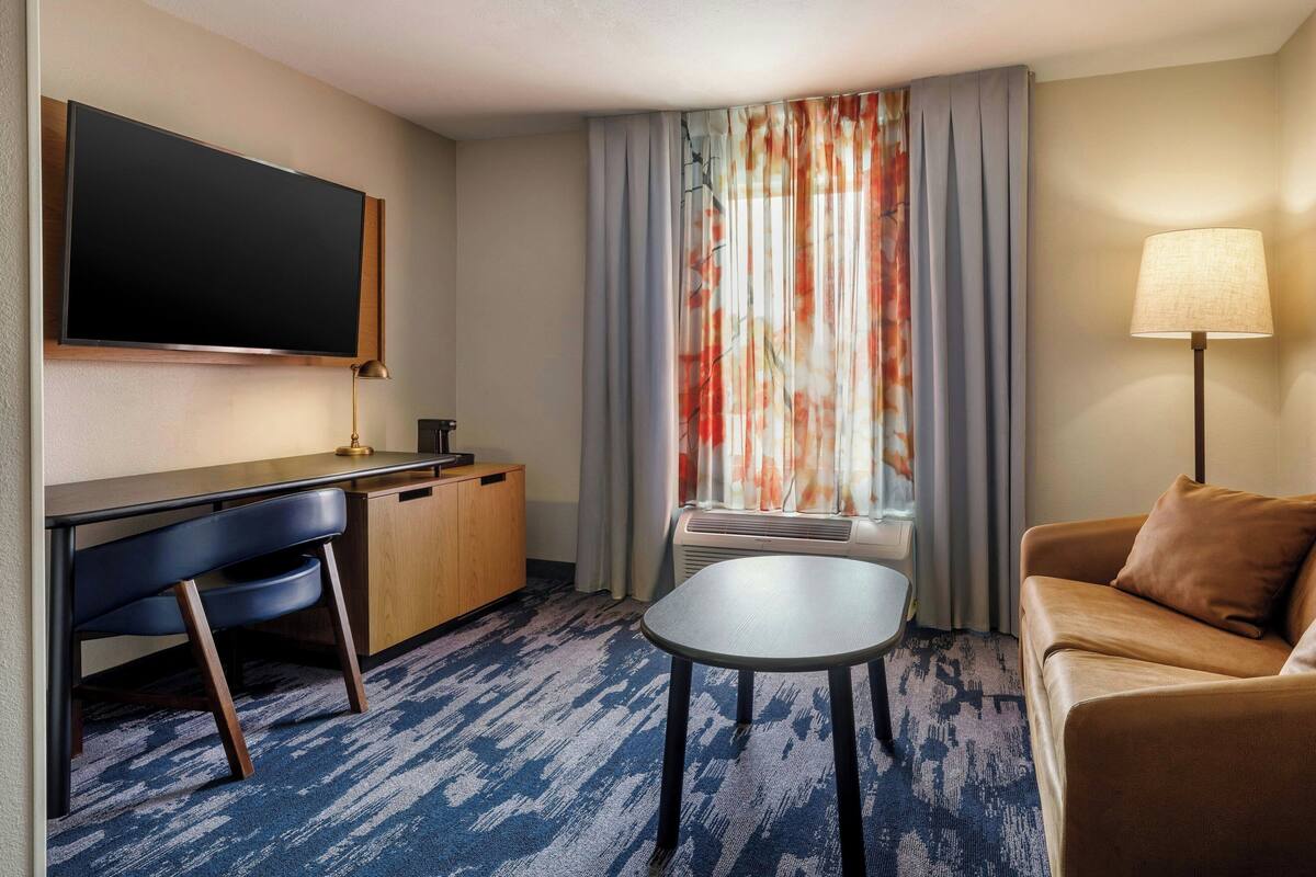 A hotel room with a flat-screen TV, a desk with a chair, a sofa, a small table, and a window with curtains.