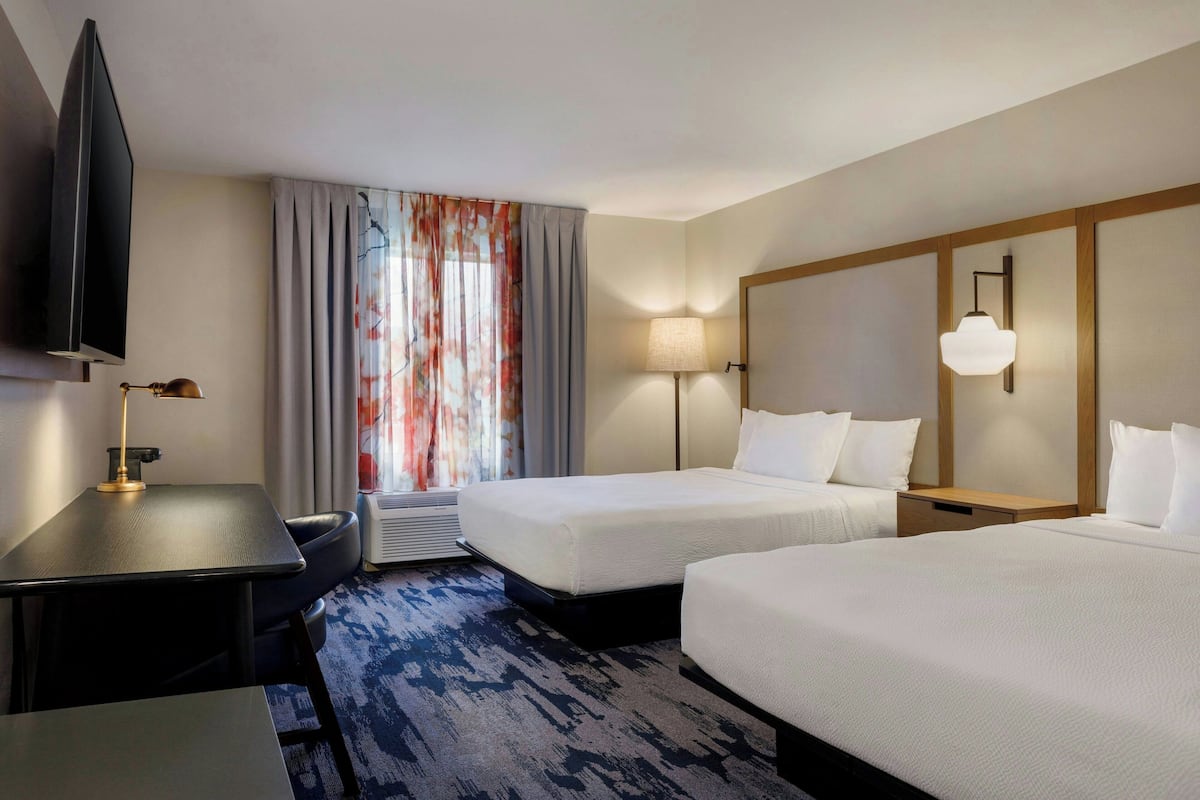 A hotel room with two beds, a desk, a TV, and a window with curtains.