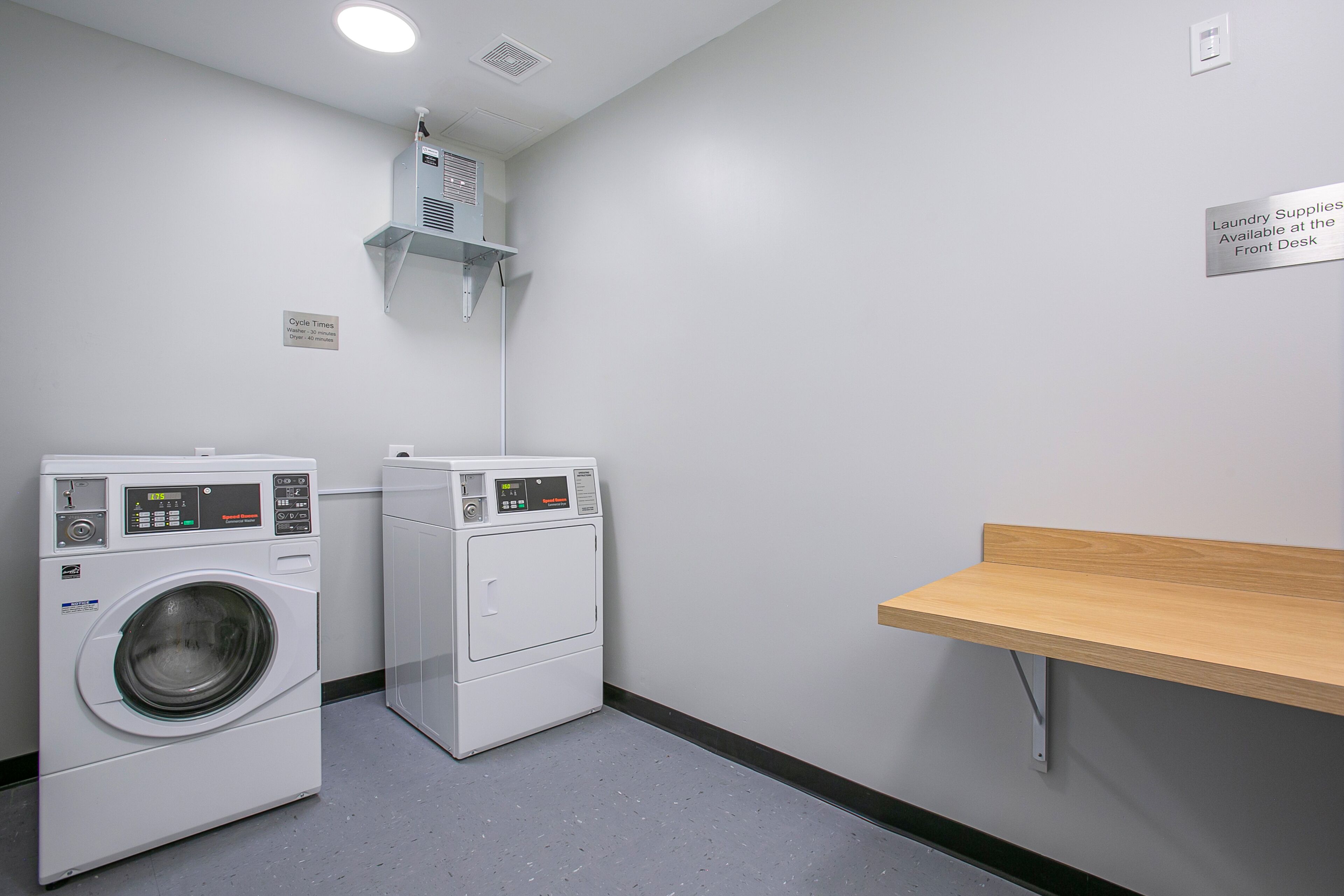 A laundry room with two front-loading washing machines, a shelf above, and a sign indicating laundry supplies are available at the front desk.
