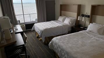 A hotel room with two beds, a desk, a chair, a lamp, and a view of the water.
