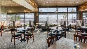 Restaurante com vista para a água, com mesas e cadeiras de madeira, janelas amplas e luminárias pendentes.