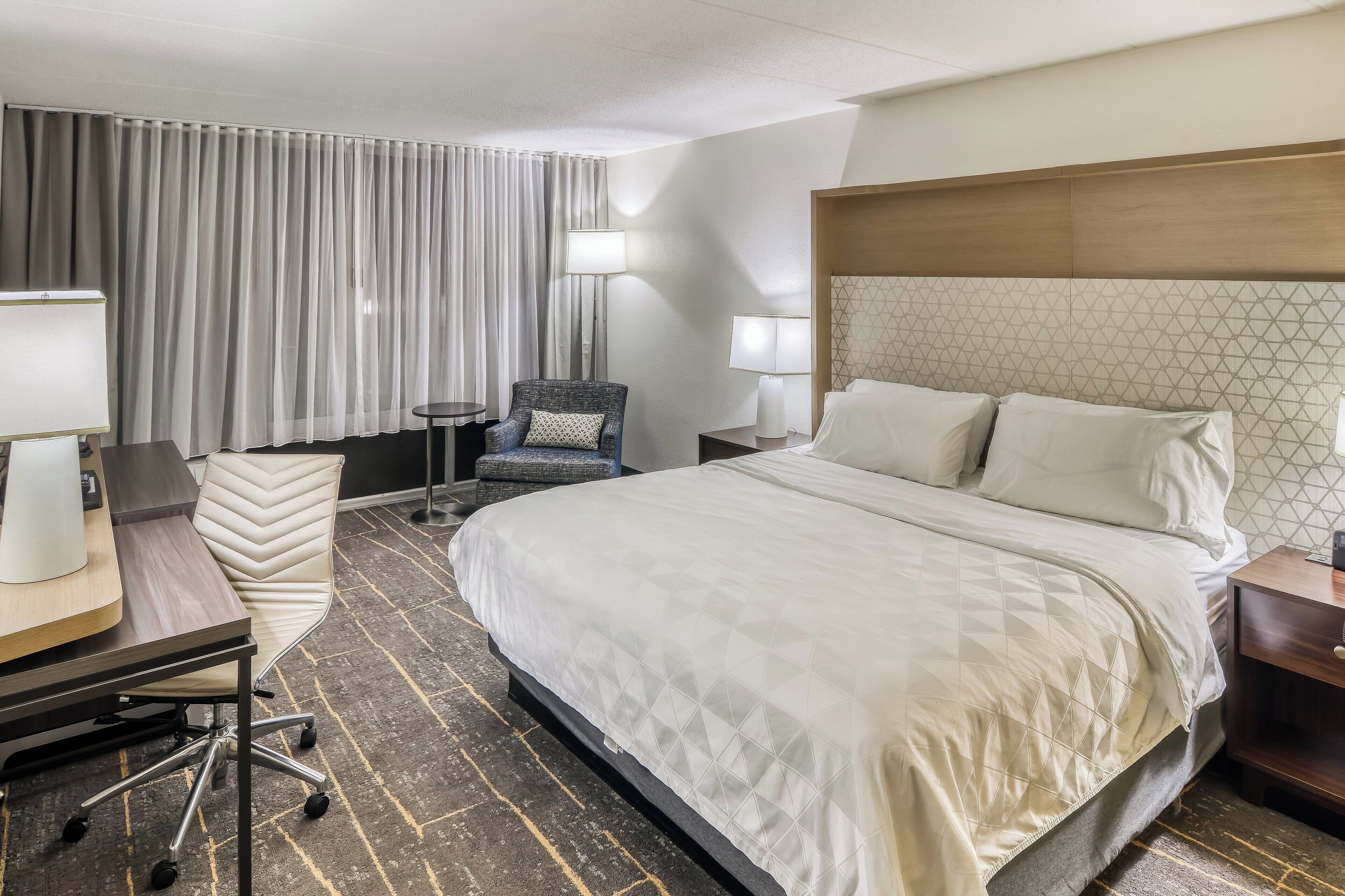 Una habitación de hotel con una cama grande, dos mesitas de noche, una silla, un escritorio con lámpara y un ventanal con cortinas.