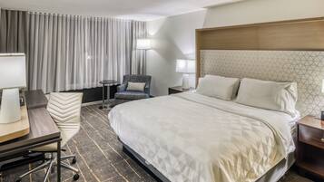 Una habitación de hotel con una cama grande, dos mesitas de noche, una silla, un escritorio con lámpara y un ventanal con cortinas.