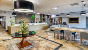 Um lobby de hotel moderno com uma mesa de recepção, uma área de refeições com uma mesa comprida e cadeiras, e uma grande luminária pendente preta.