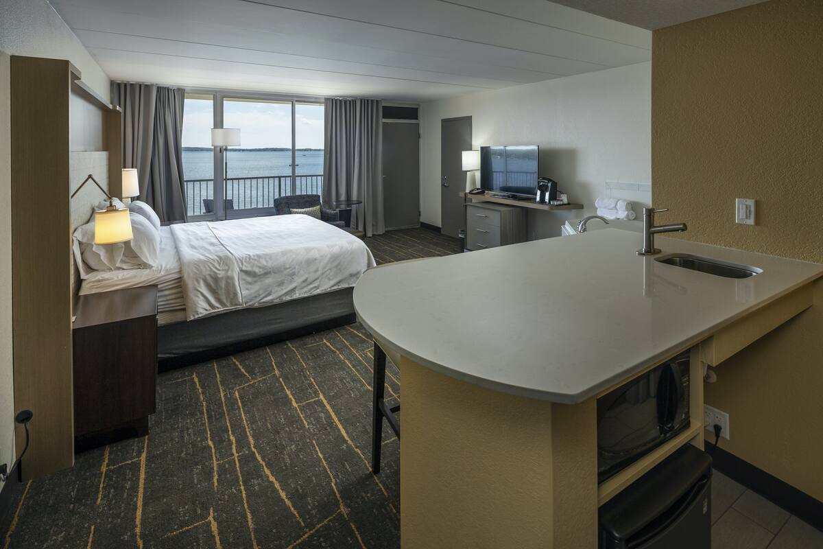 A hotel room with a bed, a desk, a TV, and a view of the sea.