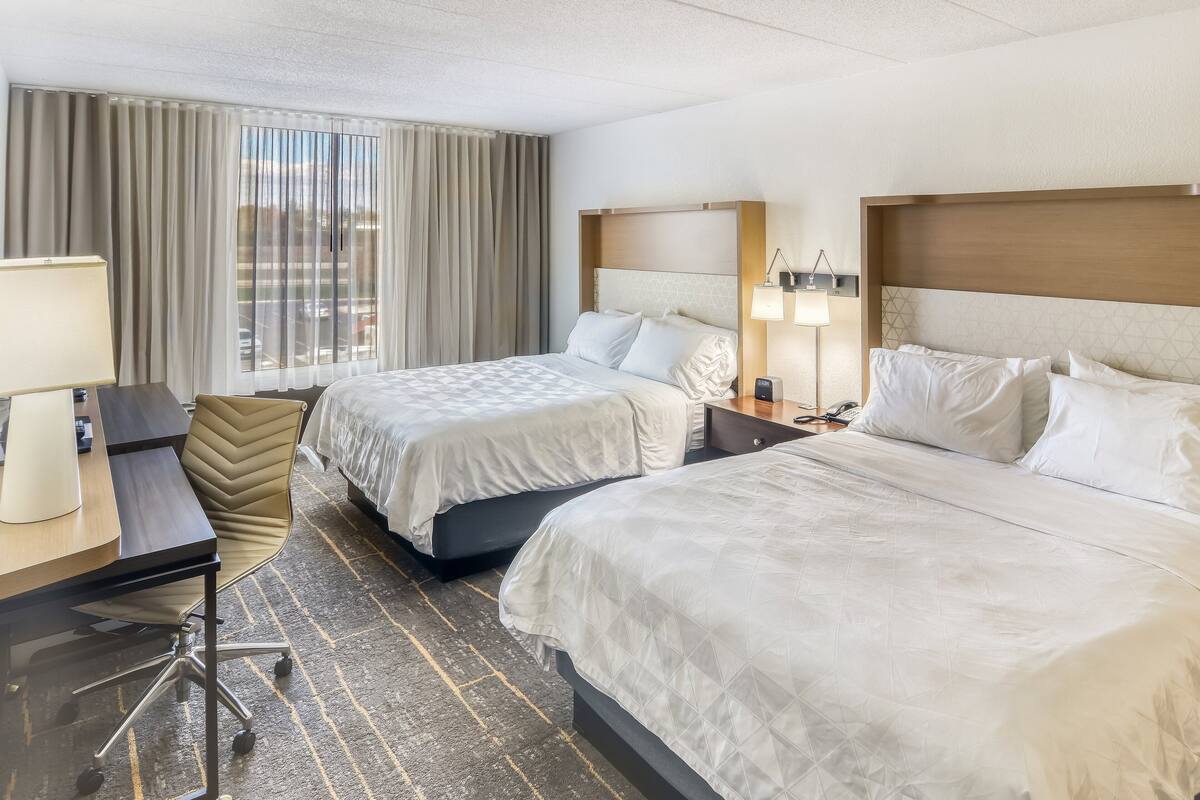 A hotel room with two beds, a desk, a chair, and a large window with curtains.