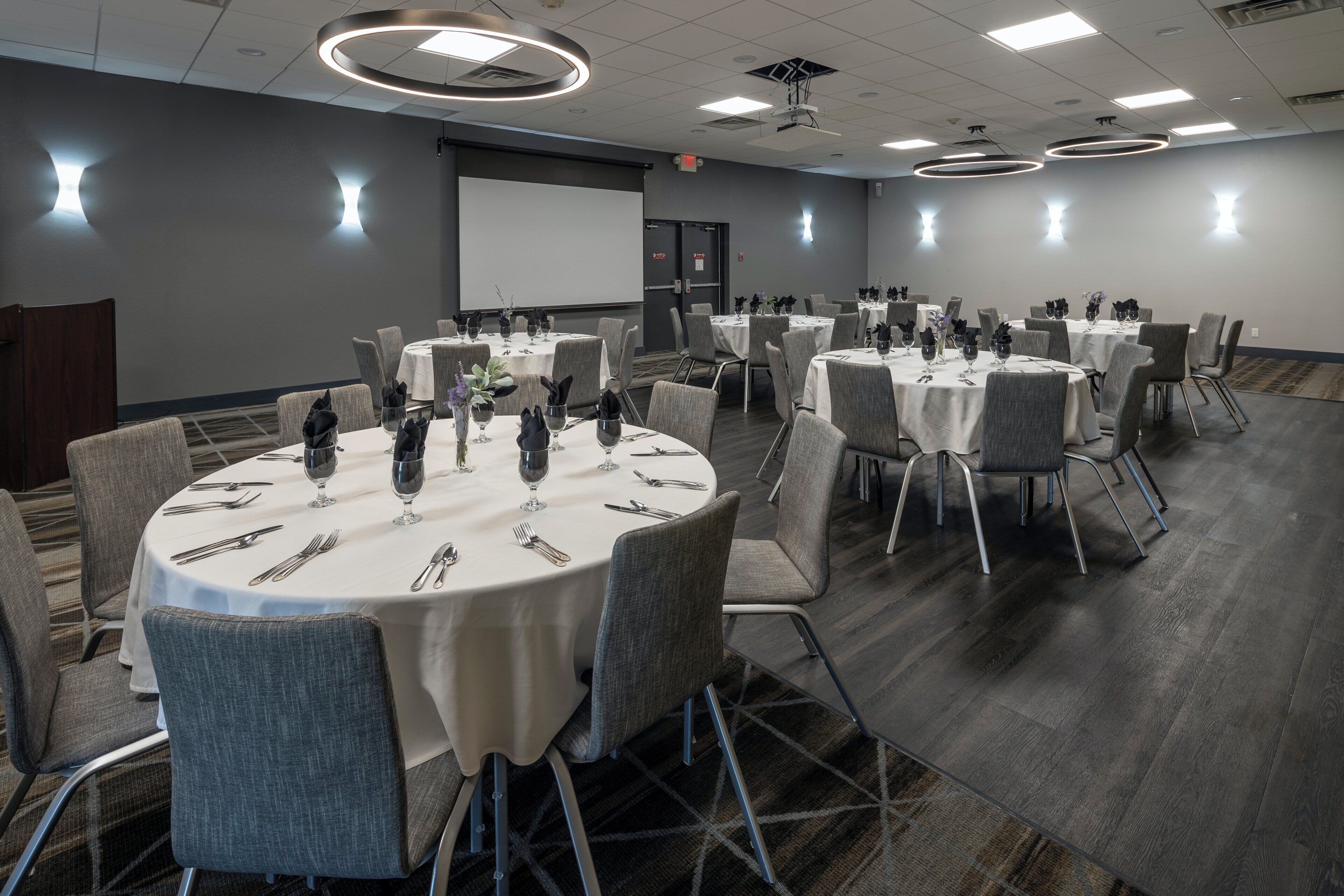 A conference room with multiple round tables, each set with white tablecloths, silverware, and wine glasses. The room has a modern design with gray chairs and a carpeted floor.