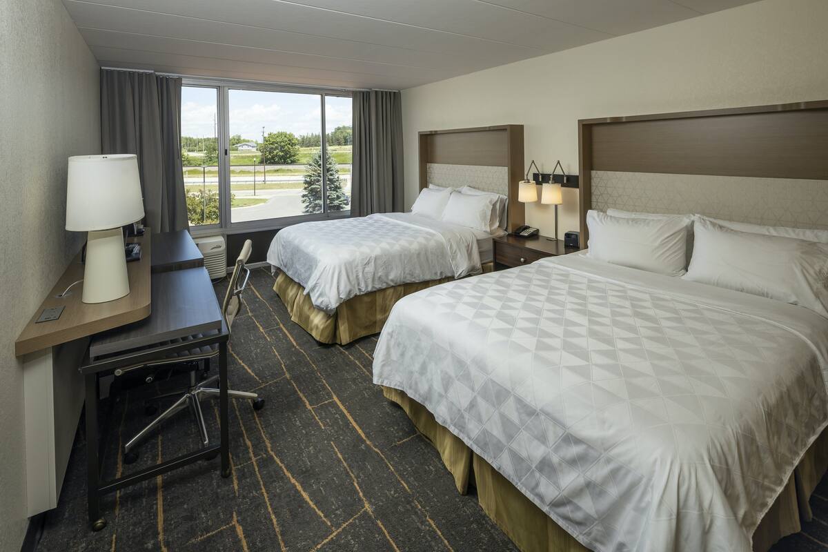 A hotel room with two beds, a desk, a chair, a lamp, and a view of the outdoors.