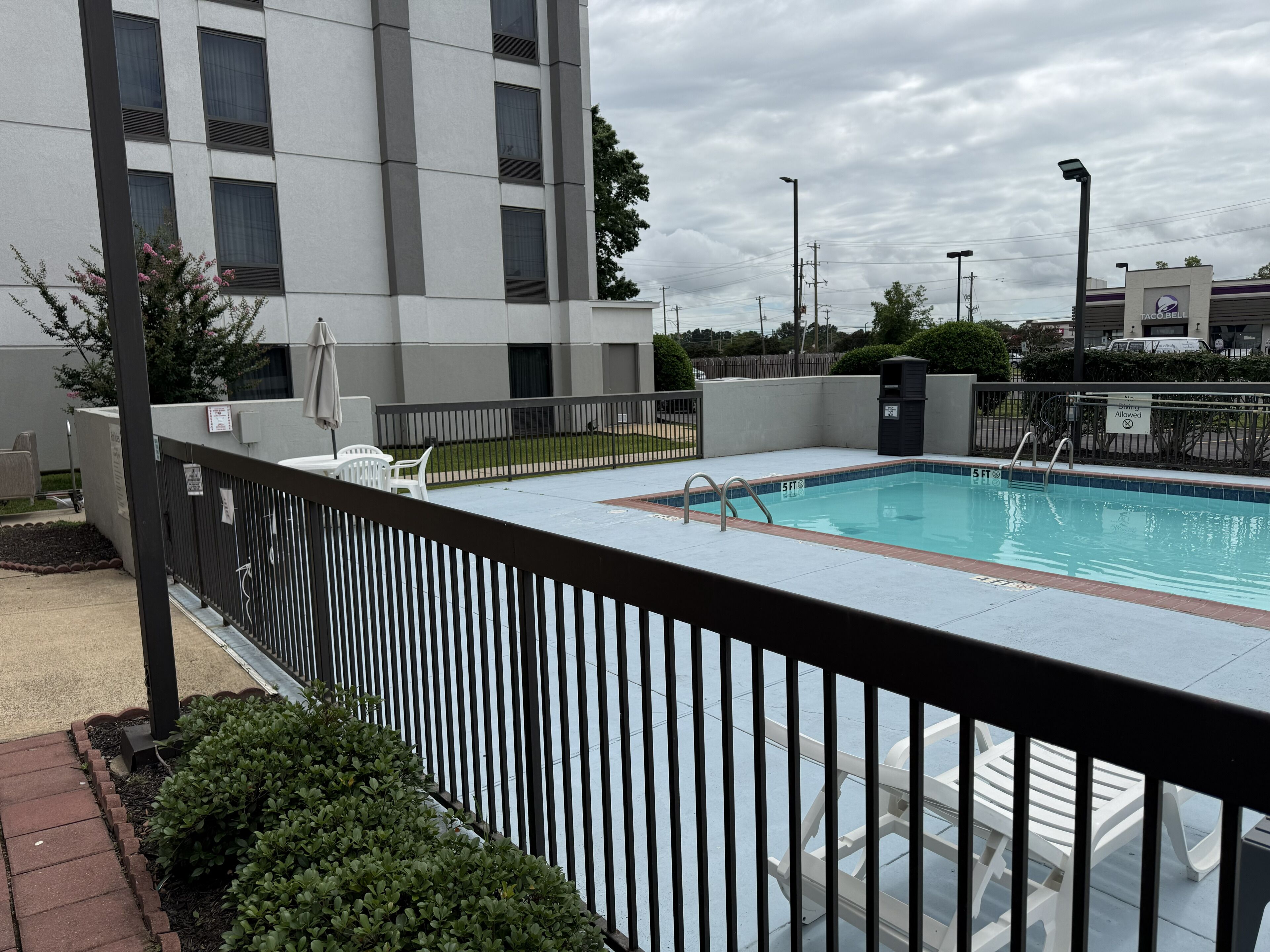 A swimming pool with a 5 ft depth marker, a fenced deck area, and a building with multiple windows in the background.