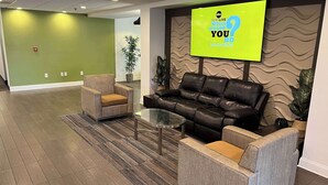 Eine Lobby mit grüner Wand, einer schwarzen Ledersofa, zwei Sesseln und einem Glastisch. Ein großer Bildschirm zeigt 'News Live World You Do Marathon'.