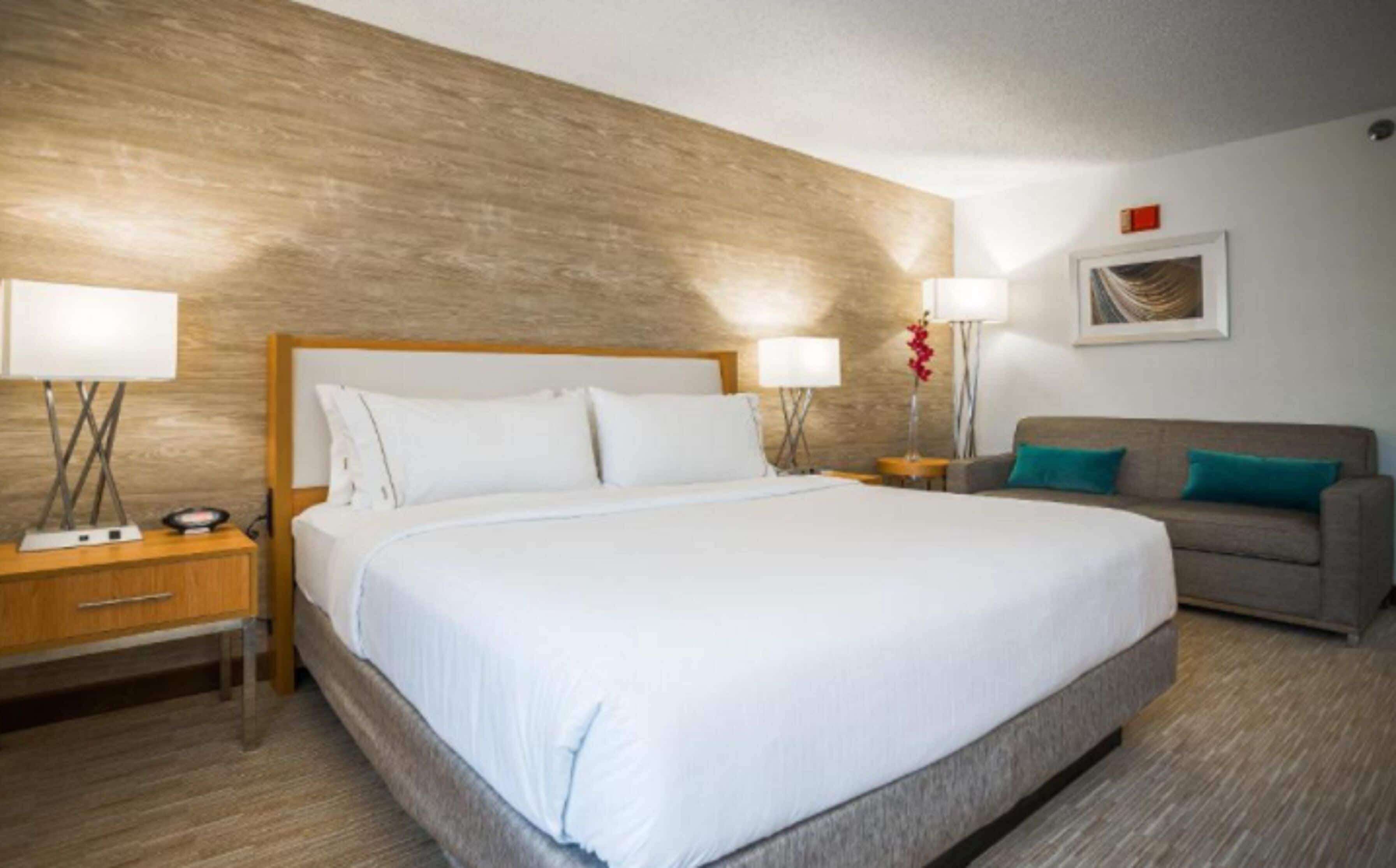 A hotel room with a large bed, a grey sofa, two bedside tables with lamps, and a wooden accent wall.