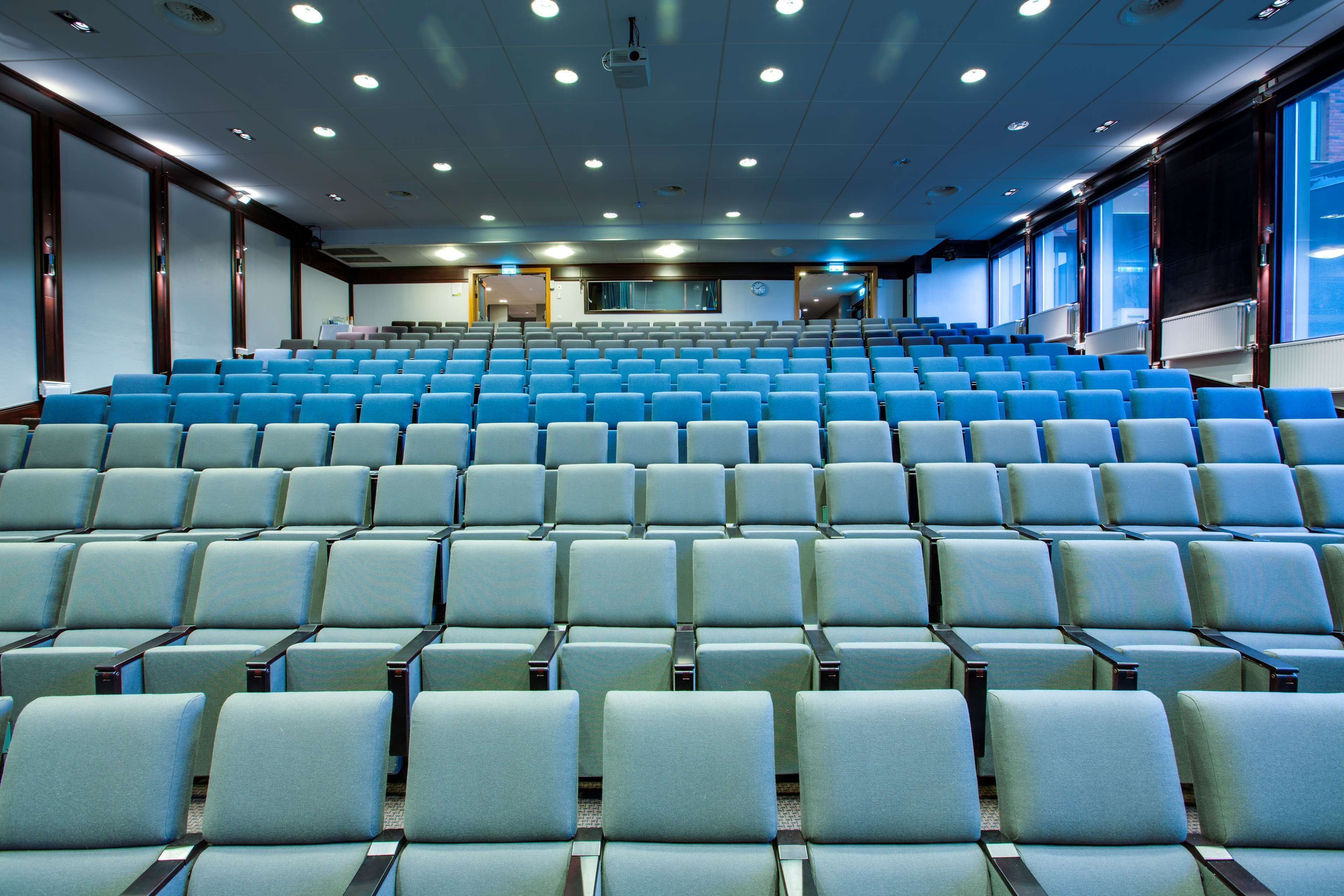Een groot, modern auditorium met rijen blauwe en witte stoelen, een podium aan de voorzijde en grote ramen langs de muren.