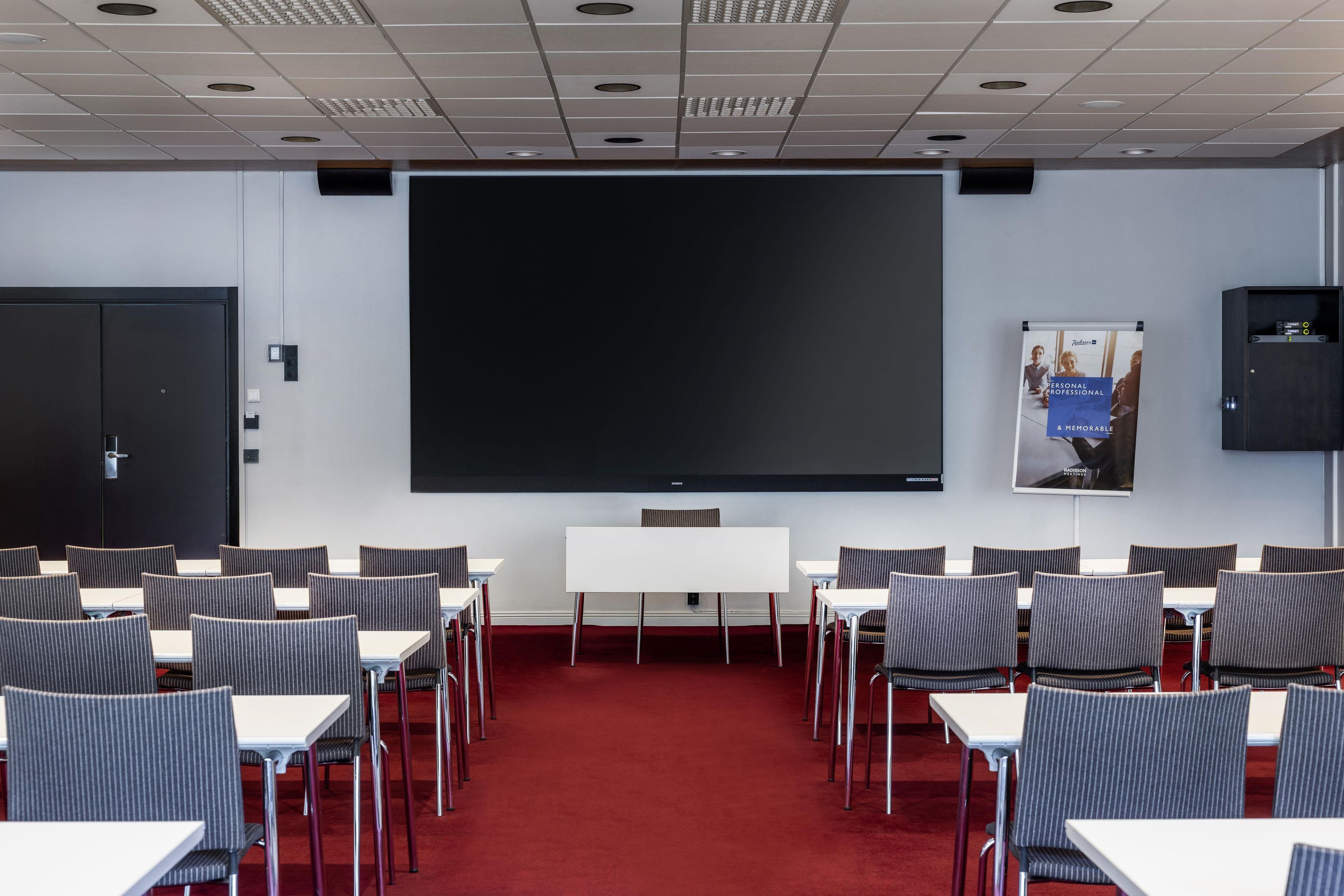 Un salón de conferencias con una pantalla grande, varias mesas y sillas dispuestas para presentaciones.