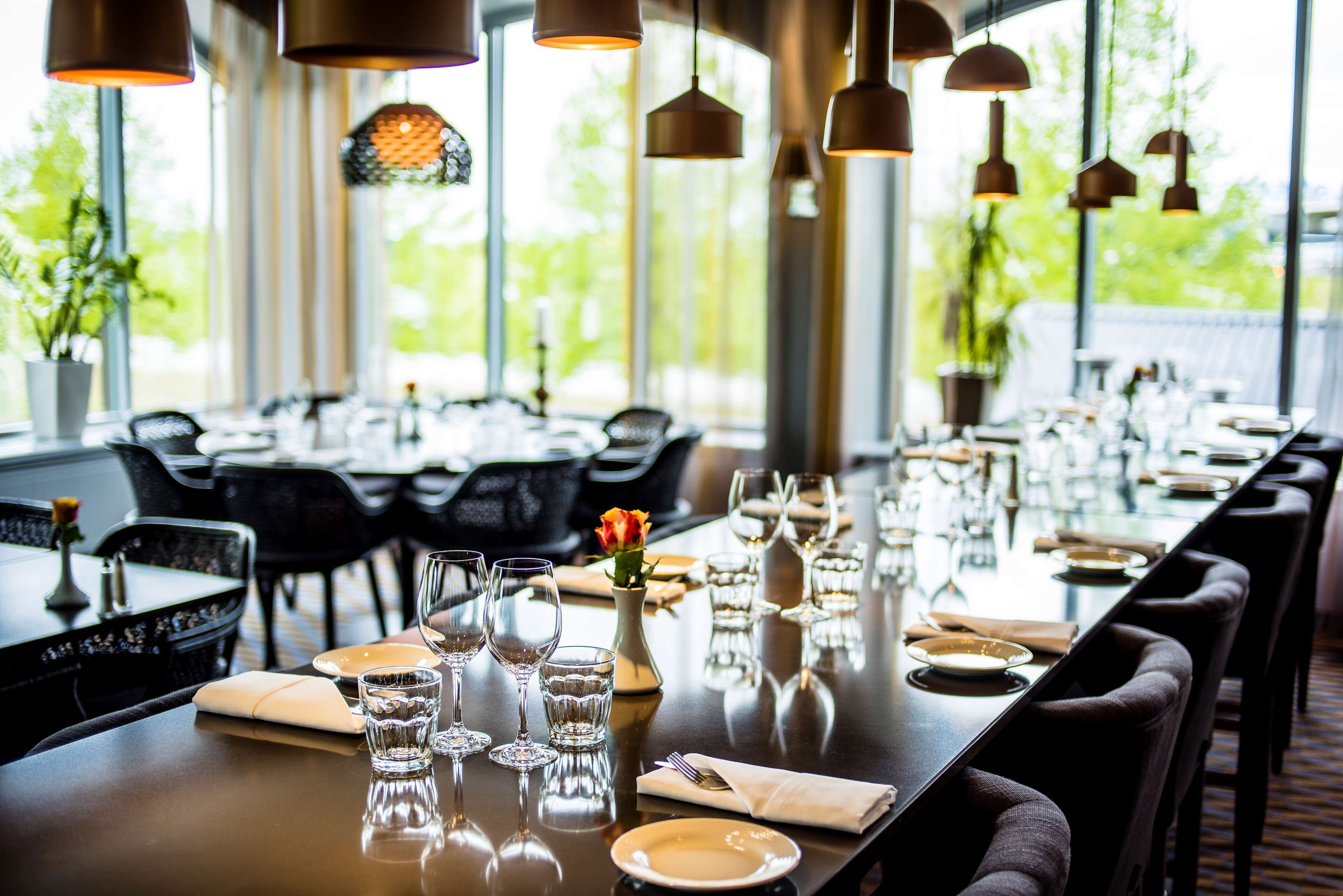 A restaurant with a long table set for dining, multiple chairs, hanging pendant lights, and a view of greenery outside.