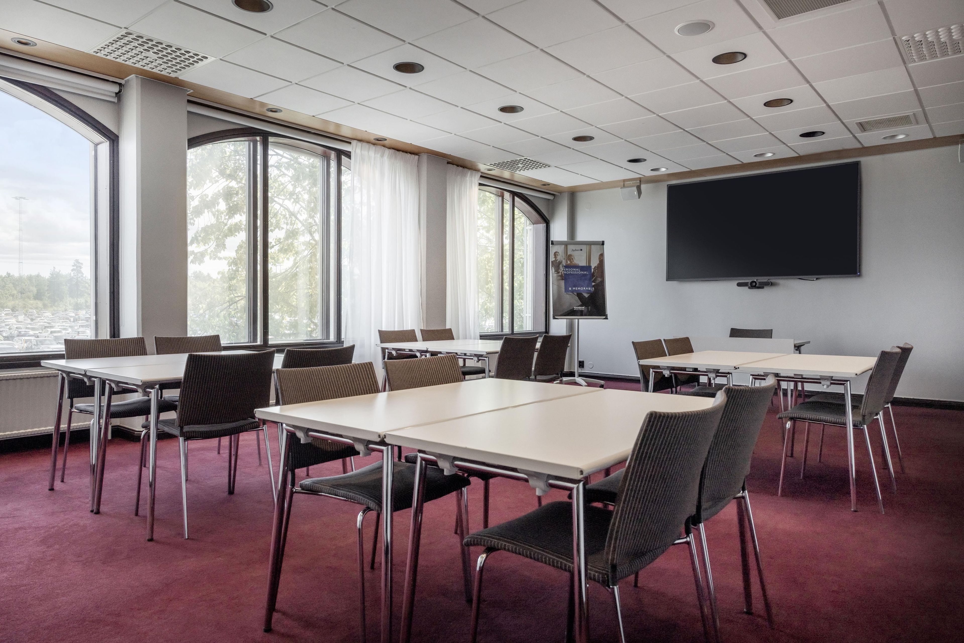 Un salón de conferencias con mesas y sillas, una pantalla grande y ventanas con cortinas.