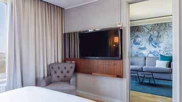 A hotel room with a large bed, a flat-screen TV mounted on a wooden cabinet, a grey armchair, and a sofa with a blue cushion.