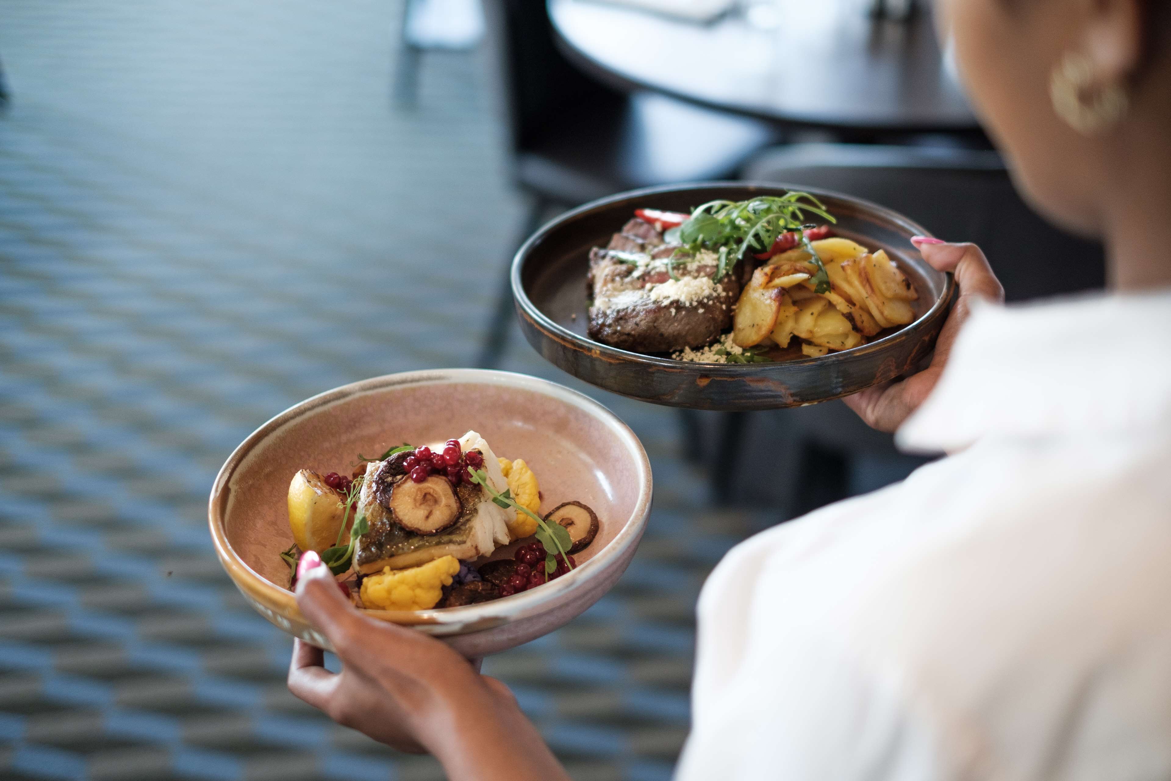 A person holding two bowls of food, one with a steak and vegetables, the other with potatoes and greens.