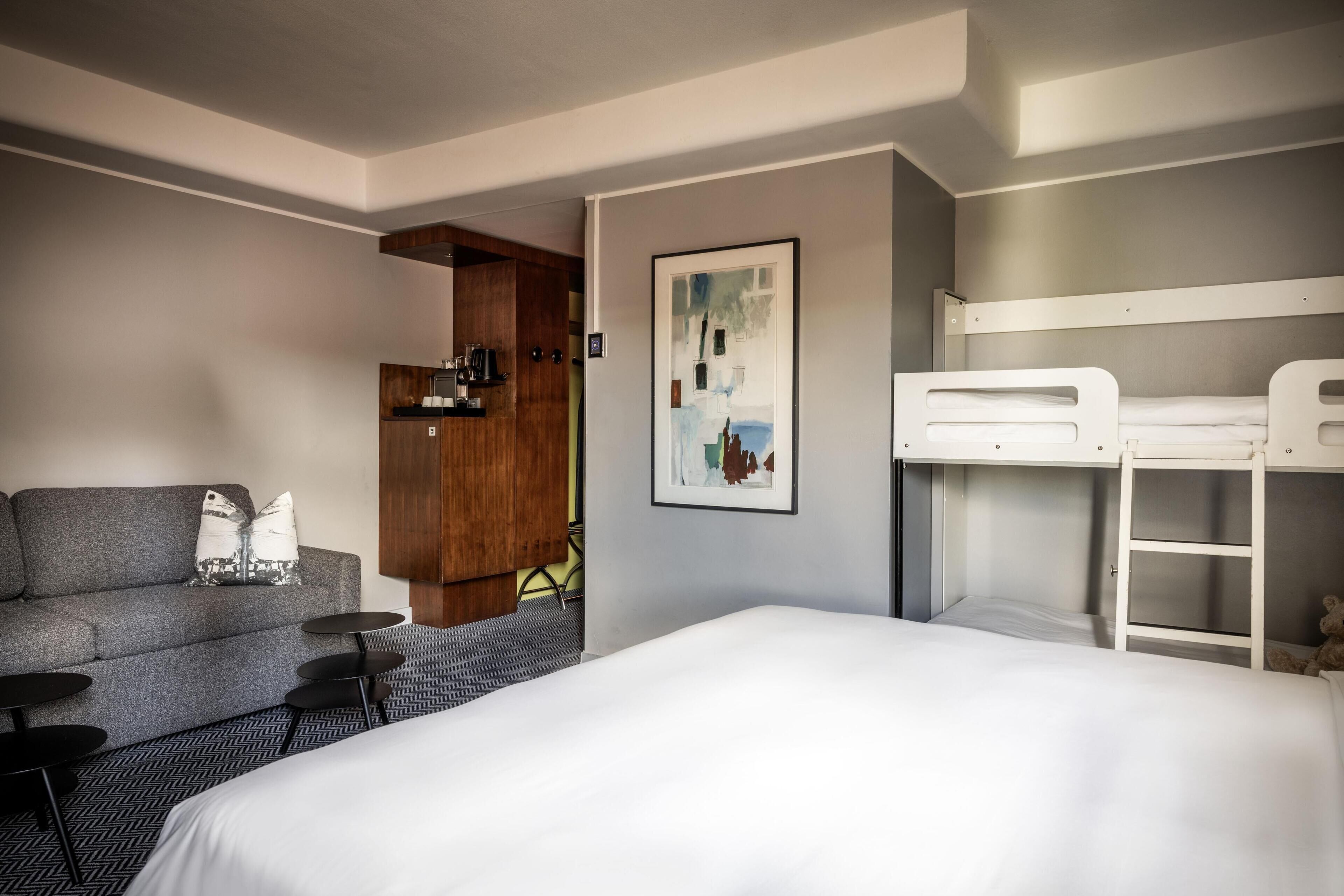 A hotel room with a bed, a sofa, a small table, a coffee machine, and a bunk bed.