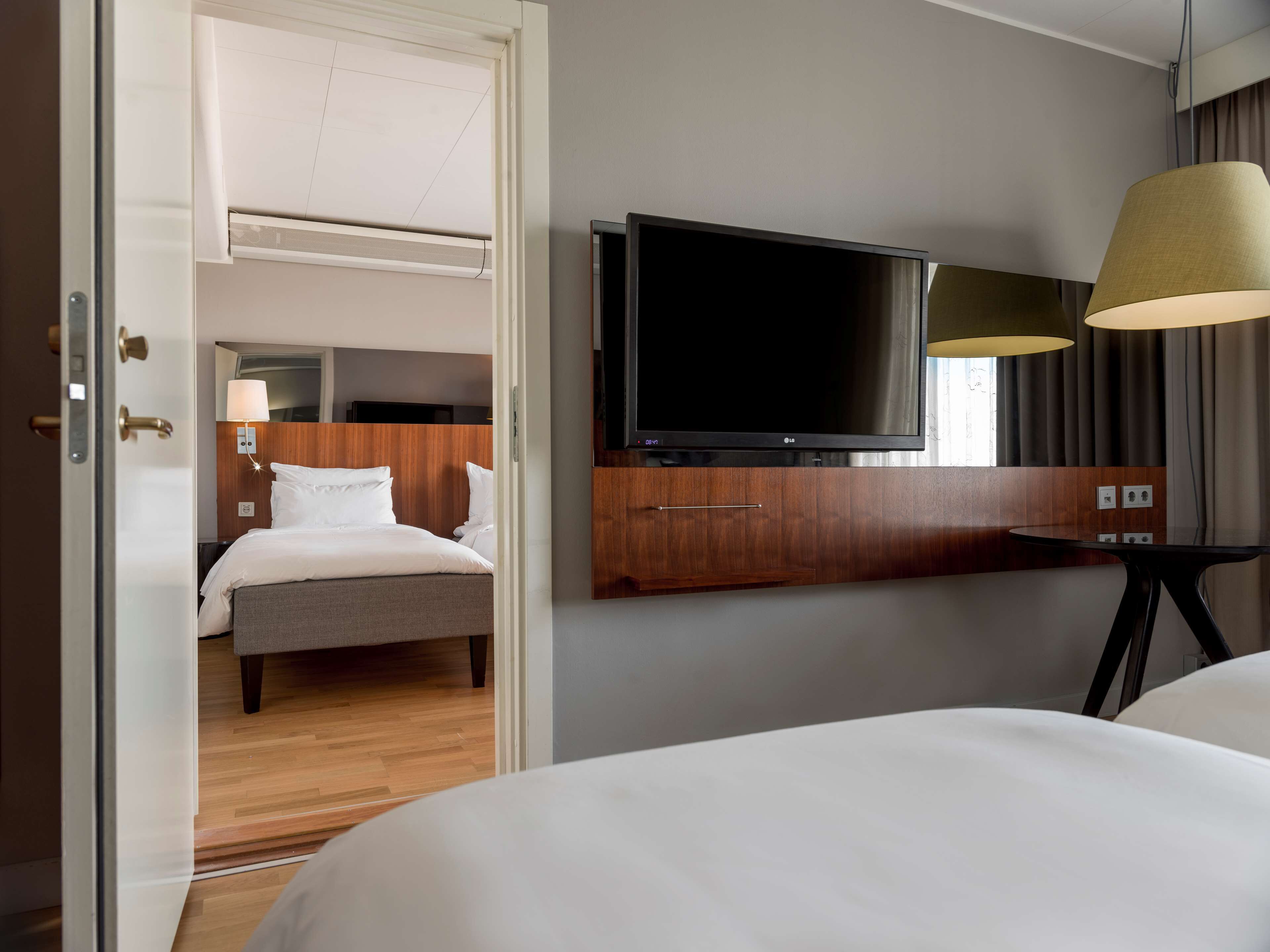 A hotel room with a bed, a TV mounted on the wall, a desk with a lamp, and a door leading to another room.