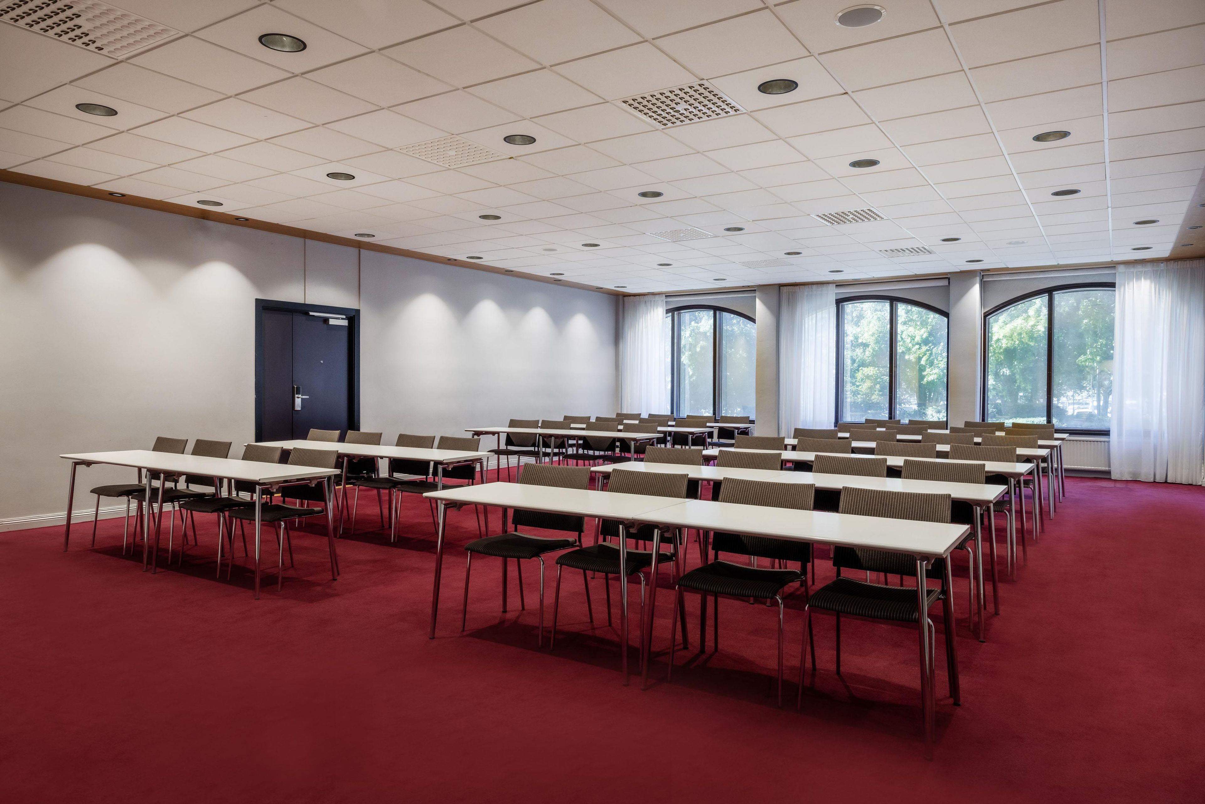Un salón de conferencias con varias mesas y sillas, amplias ventanas y un suelo cubierto de moqueta roja.