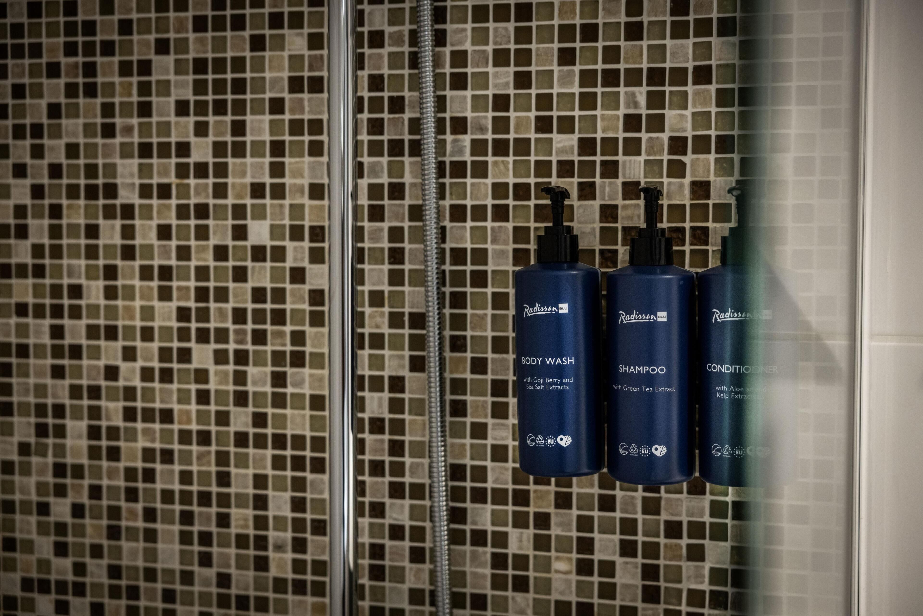 Un bagno con una parete piastrellata e tre flaconi di prodotti da bagno Radisson: bagnoschiuma, shampoo e balsamo.