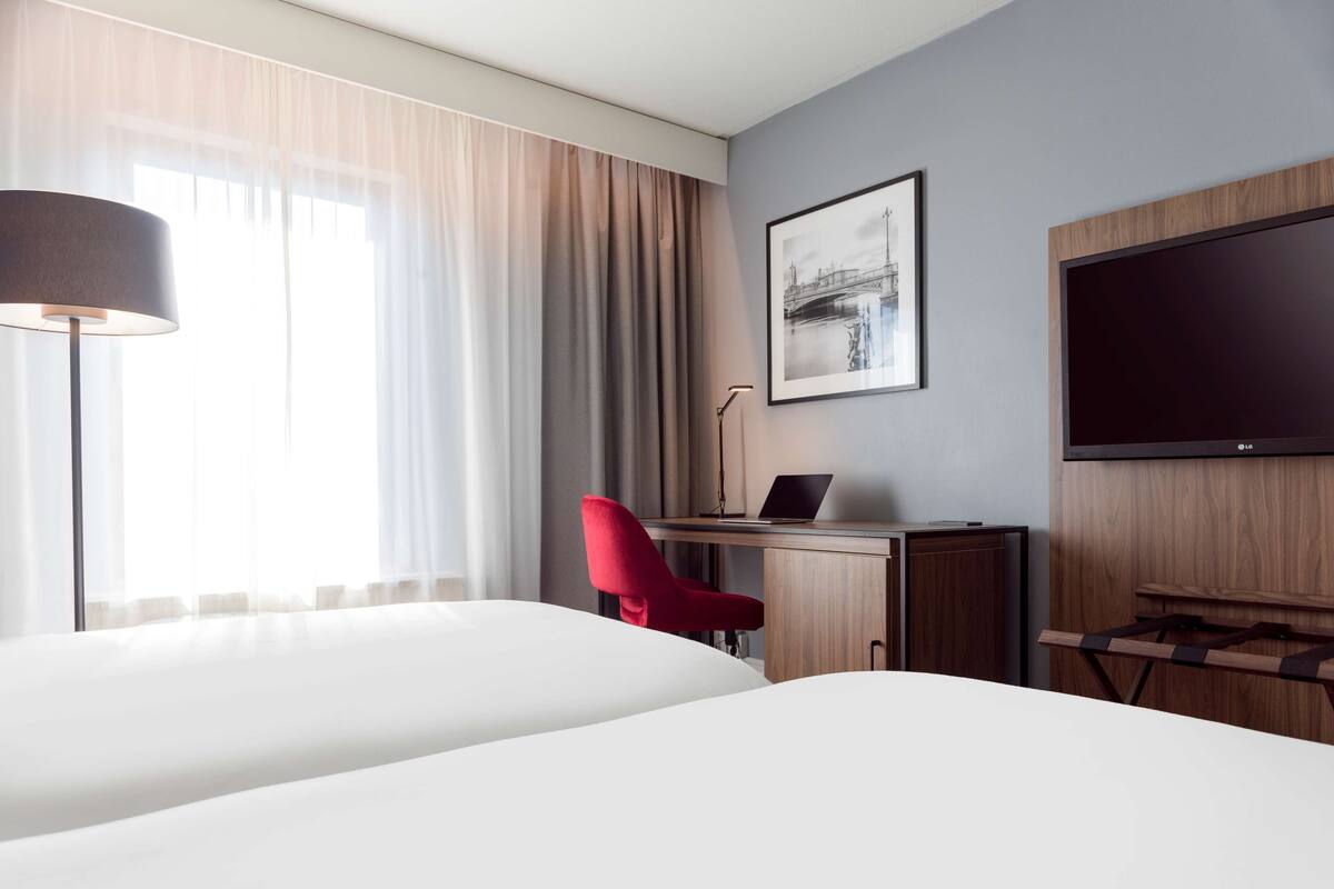 Une chambre d’hôtel avec un lit, un bureau avec un ordinateur portable, une chaise rouge, une télévision et un tableau encadré au mur.