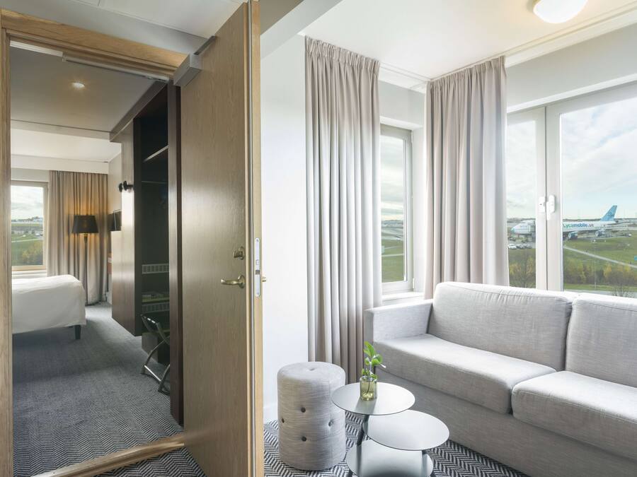 Habitación de hotel con cama, sofá, una mesita y vistas al aeropuerto.
