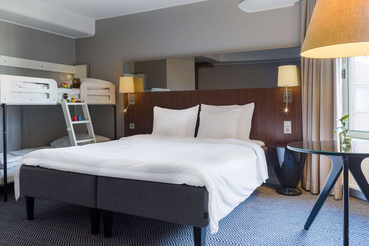 Une chambre d’hôtel avec un lit superposé, un bureau avec une lampe, une petite table avec un vase et un tapis à motifs.