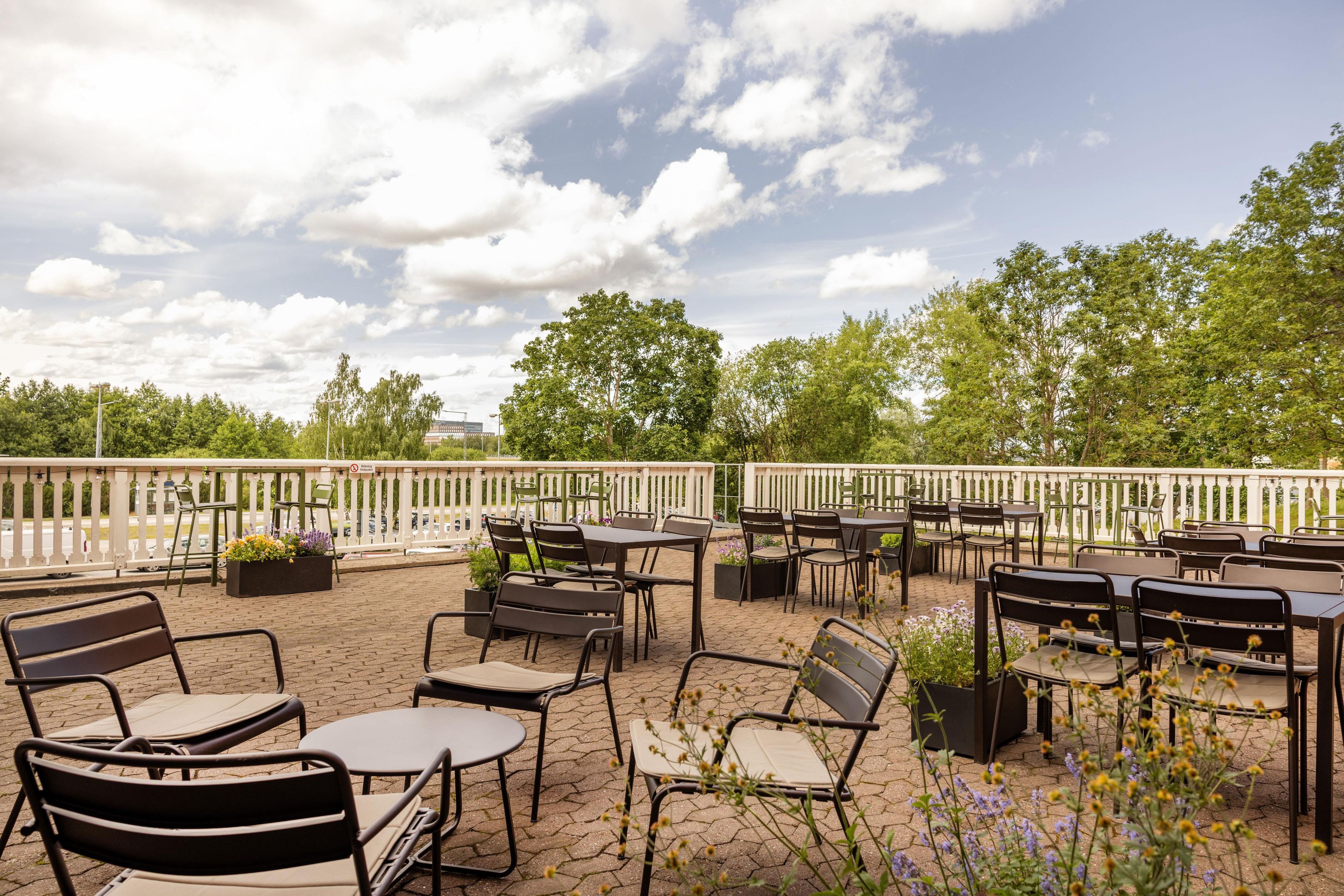 Un espace extérieur aménagé en terrasse, avec des tables et des chaises, entouré d’une clôture blanche, et offrant une vue sur des arbres et un ciel nuageux.