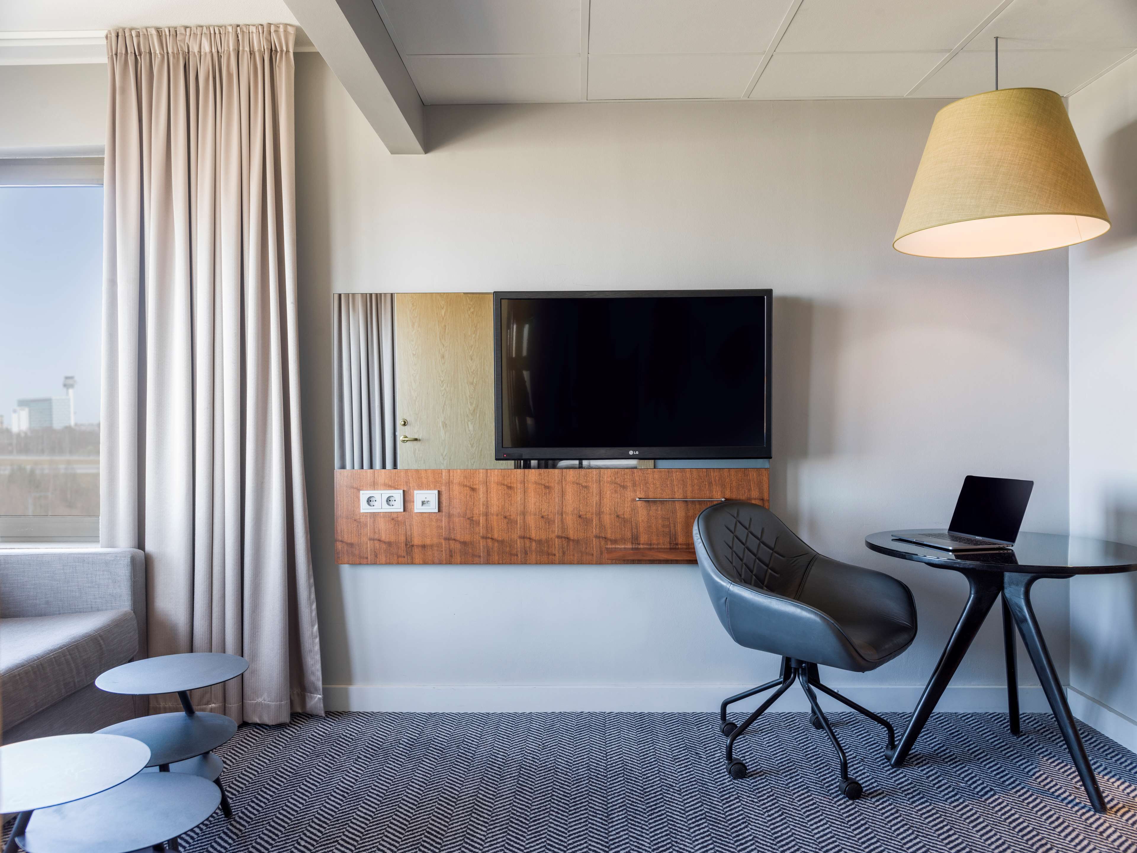 A hotel room with a flat-screen TV, a laptop on a small table, a grey office chair, and a large window with curtains.