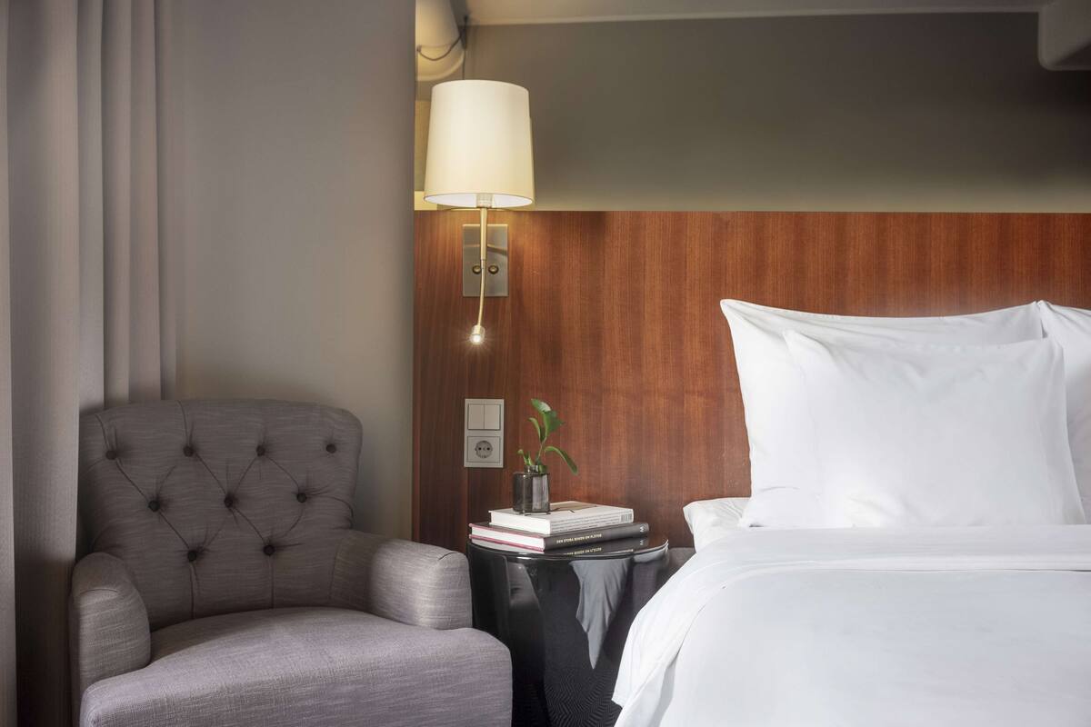 Une chambre d’hôtel avec un fauteuil à capitons, une table de chevet avec une lampe, une plante et une pile de livres.