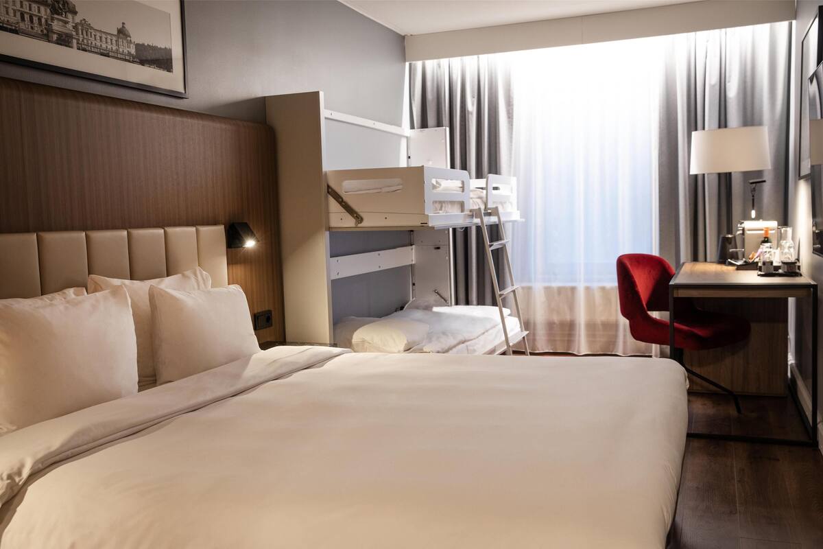 Une chambre d’hôtel avec un grand lit, un bureau avec une chaise, une échelle et une fenêtre avec des rideaux.
