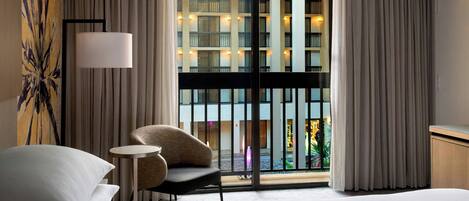 A hotel room with a large bed, a chair, a small table, a lamp, and a view of a building with balconies and lights.