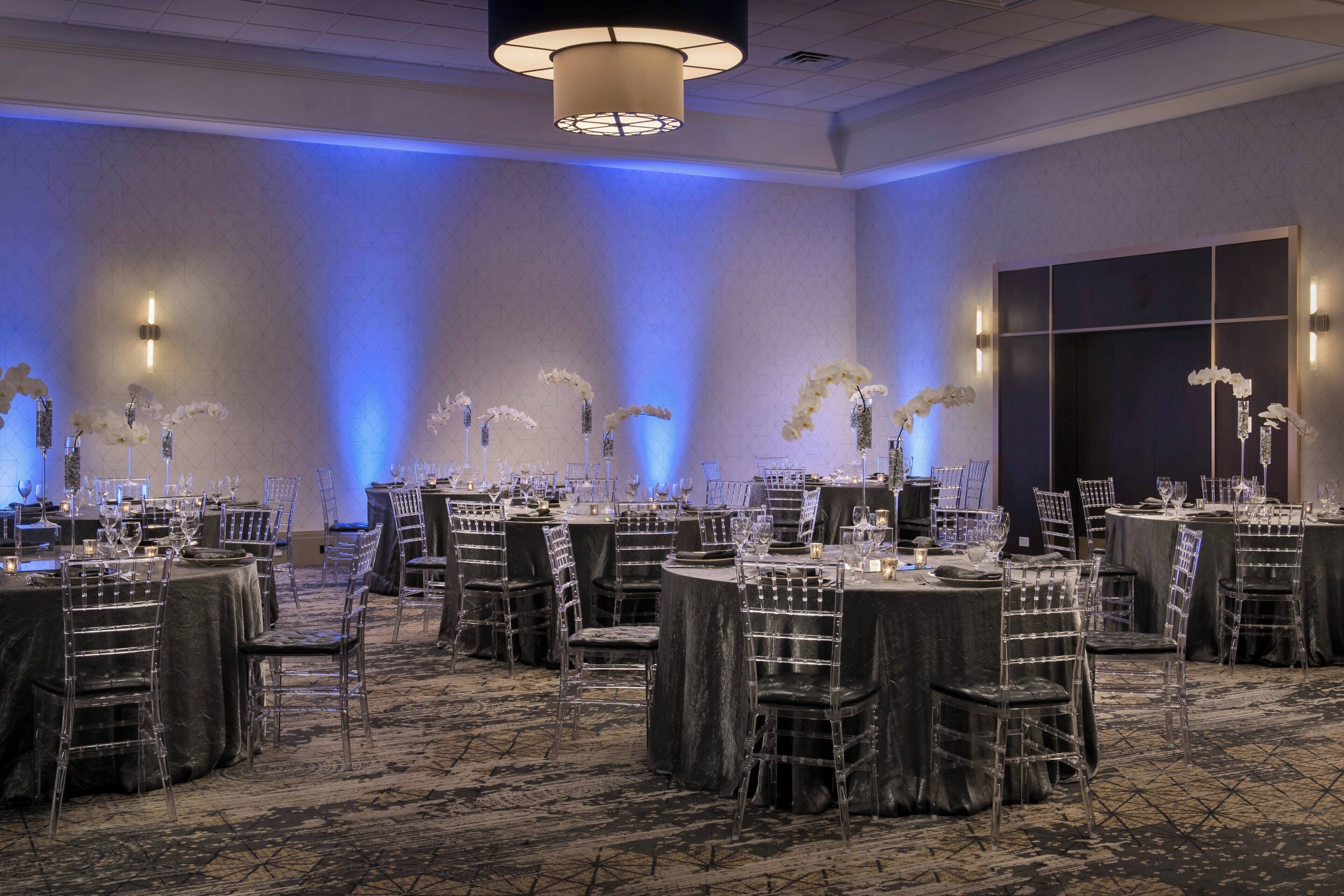 A banquet hall with round tables, clear chairs, and floral centerpieces.