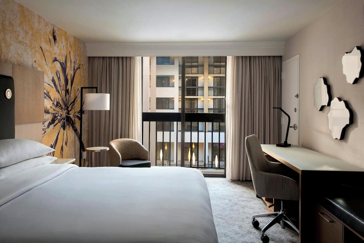 A hotel room with a large bed, a desk, a chair, and a view of a building through the window.