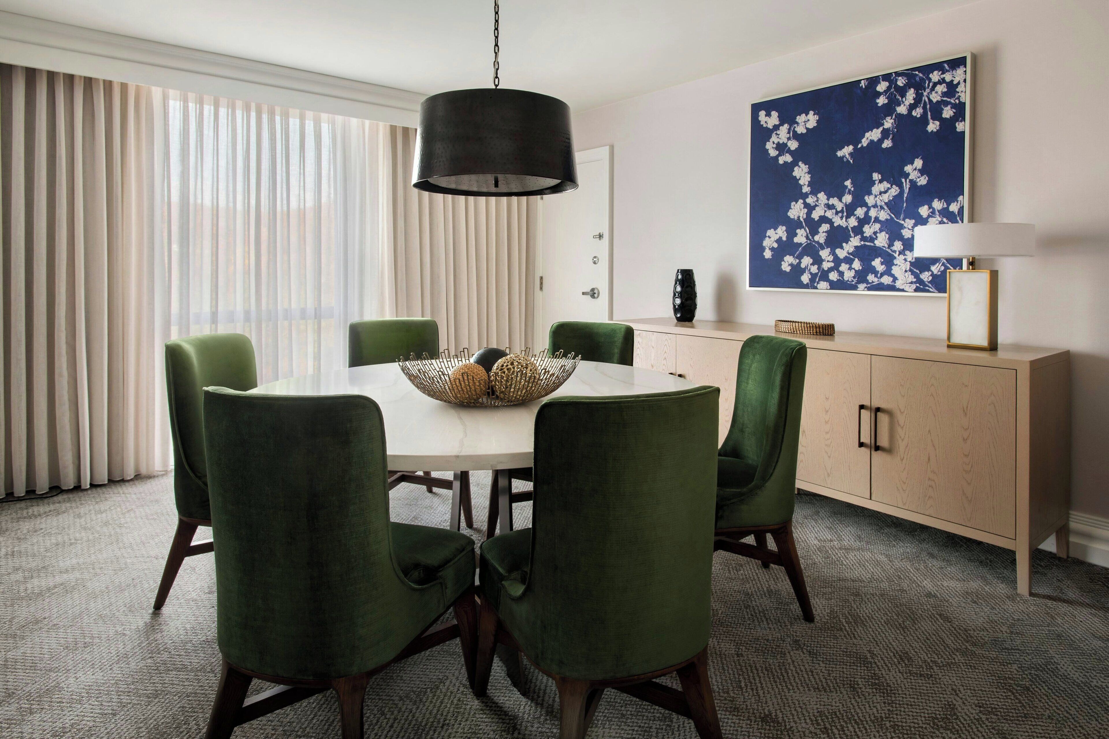 Un comedor con una mesa redonda, seis sillas verdes tapizadas, una lámpara colgante negra, un florero blanco y una obra de arte floral azul en la pared.