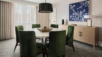 Un comedor con una mesa redonda, seis sillas verdes tapizadas, una lámpara colgante negra, un florero blanco y una obra de arte floral azul en la pared.