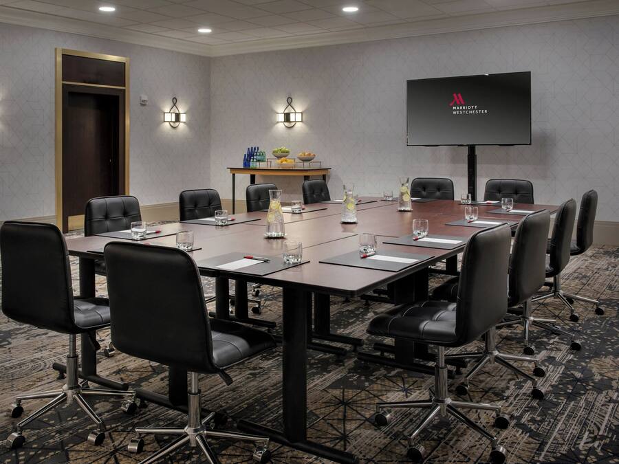 A conference room with a long table, black chairs, a screen, and a small table with fruits and condiments.