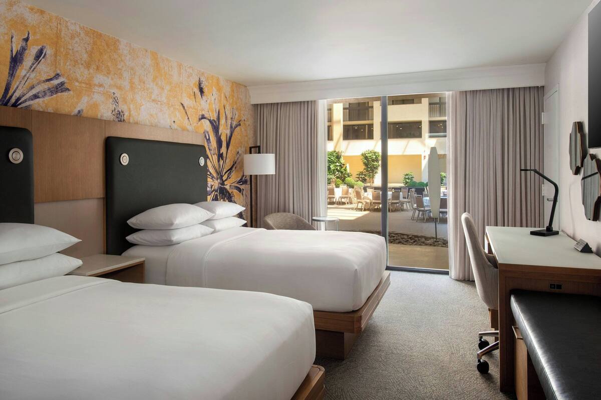 A hotel room with two beds, a desk, and a view of the outdoor patio.