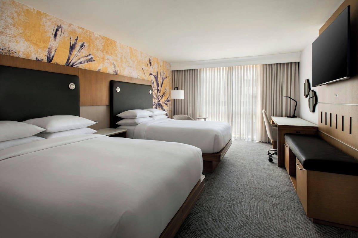 A hotel room with two beds, a desk, a chair, a TV, and a large window with curtains.