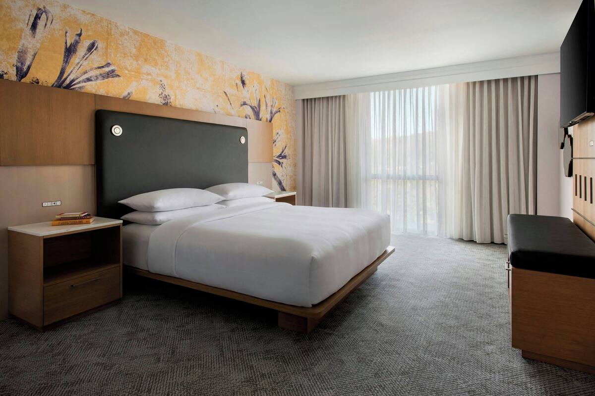 A hotel room with a bed, a nightstand, a desk, and a large window with curtains.